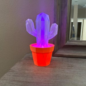 May include: A geometric cactus-shaped lamp in an orange pot. The cactus glows with purple and white light, while the pot emits a red glow. The lamp is on a wooden surface.