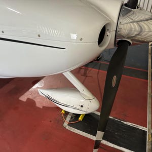 May include: Close-up of a white airplane fuselage with a black propeller. The propeller has a silver spinner and a black blade. The wing has a white and black design. The background is a red floor.