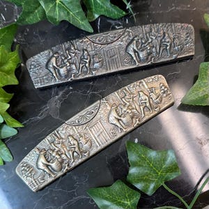 May include: Two rectangular metal hair barrettes featuring a dark, antiqued silver finish. Each barrette displays a detailed, embossed relief scene of figures working with barrels in an outdoor setting, accented by a decorative floral motif.