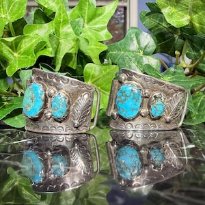 May include: Two silver-toned belt buckles with turquoise stones. Each buckle features three turquoise stones and a leaf design. The buckles are displayed on a reflective surface, with green foliage in the background.