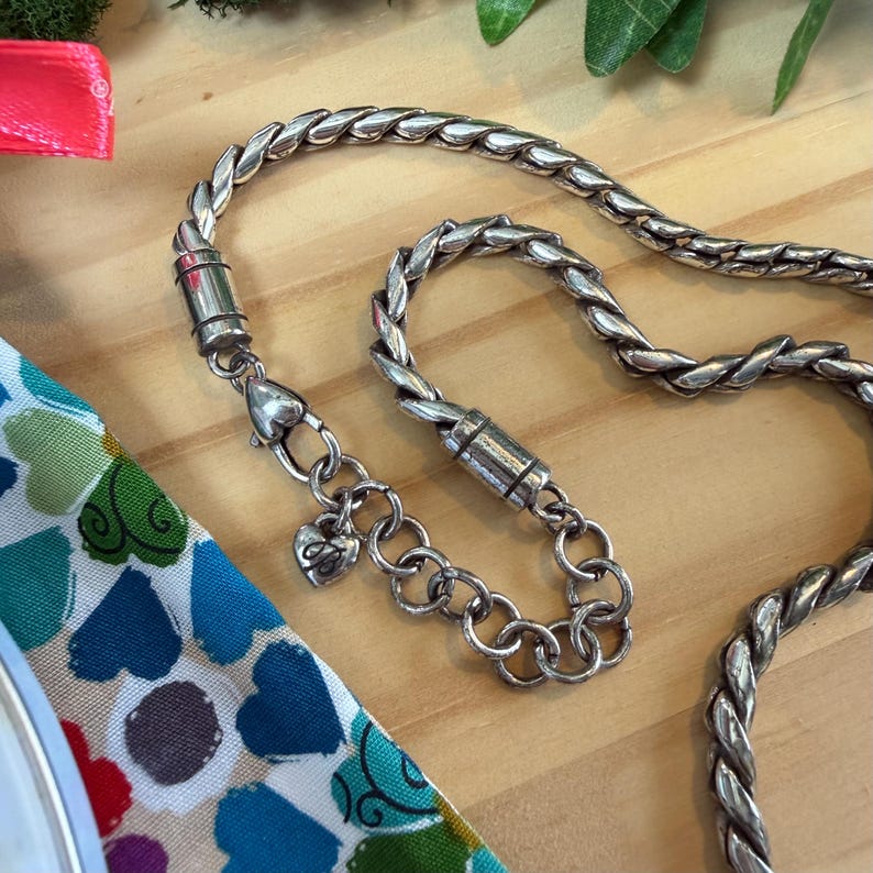 May include: A silver-toned necklace with a braided chain design. The necklace features a heart-shaped charm and a magnetic clasp. The jewelry is displayed on a wooden surface with a colorful patterned fabric in the background.