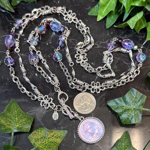 May include: A silver-toned necklace with multiple strands, featuring iridescent purple beads and a pendant with a cameo design. The necklace is displayed on a black surface with green leaf accents. A U.S. quarter is next to the pendant.