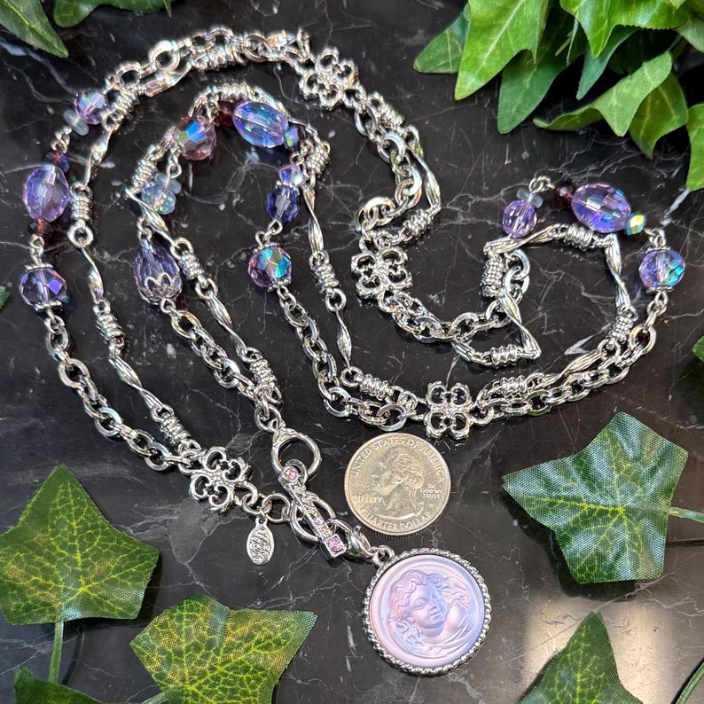 May include: A silver-toned necklace with multiple strands, featuring iridescent purple beads and a pendant with a cameo design. The necklace is displayed on a black surface with green leaf accents. A U.S. quarter is next to the pendant.