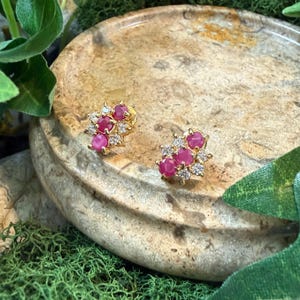 May include: A pair of gold-colored stud earrings. Each earring features a cluster of small, round, bright pink gemstones surrounded by smaller, clear gemstones. The earrings are set against a natural stone surface.