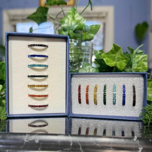 May include: A collection of colorful gemstone rings displayed in blue boxes. The rings feature a variety of colors, including red, orange, yellow, green, teal, blue, and black. The rings are set in silver bands.