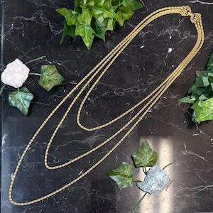 May include: A long, gold-toned chain necklace featuring a delicate, repeating link pattern. The necklace is designed with a multi-layered appearance, showcasing a continuous, slender gold chain that loops to create two distinct, elegant strands.