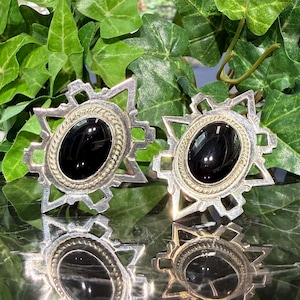 May include: A pair of silver-toned earrings featuring a central oval black stone surrounded by a twisted rope border. The setting has a geometric, star-like frame with pointed edges and cut-out details, reflecting a polished metallic finish.