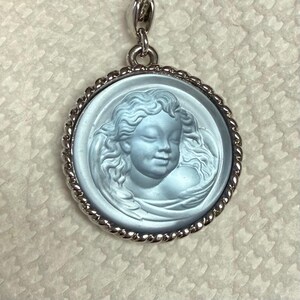May include: A round, light blue pendant with a detailed relief of a cherub's face. The cherub has curly hair and a serene expression. The pendant is set in a silver-colored, twisted rope-style frame and hangs from a matching chain.