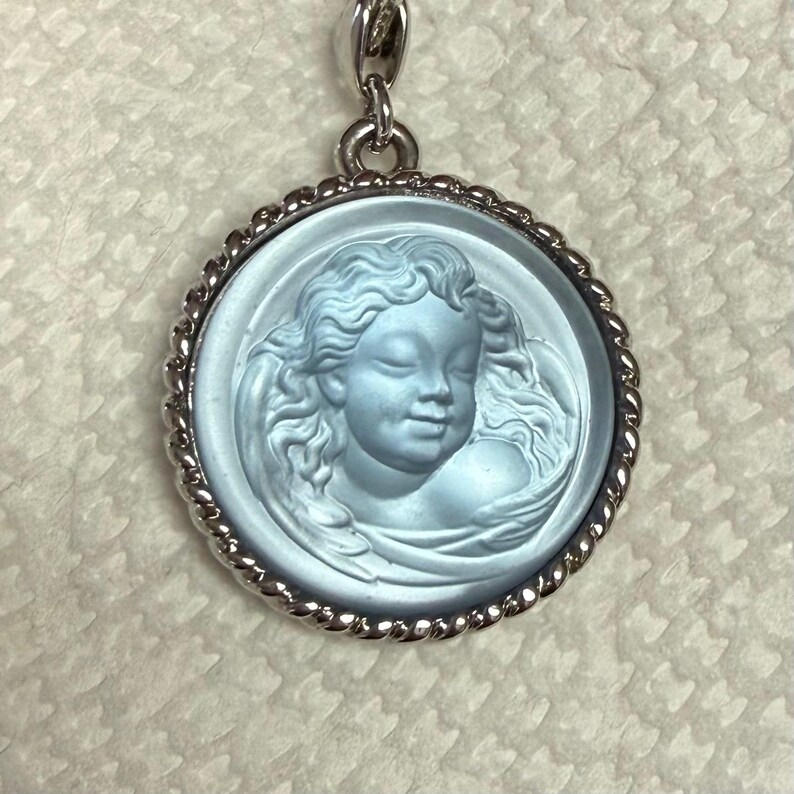 May include: A round, light blue pendant with a detailed relief of a cherub's face. The cherub has curly hair and a serene expression. The pendant is set in a silver-colored, twisted rope-style frame and hangs from a matching chain.