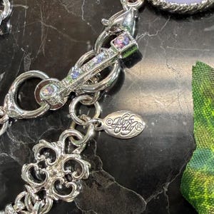 May include: Close-up of a silver-toned necklace clasp with a decorative design. The clasp features small, colorful gemstones and is connected to a chain with an ornate pendant. The brand name "Kirk's Folly" is visible on a small tag.