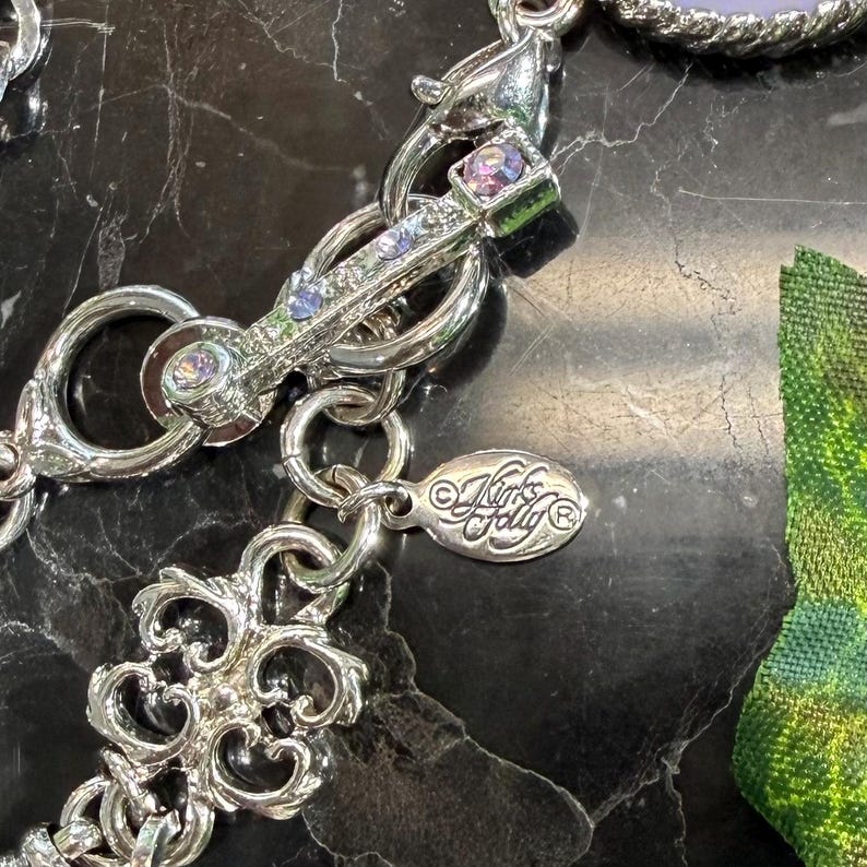 May include: Close-up of a silver-toned necklace clasp with a decorative design. The clasp features small, colorful gemstones and is connected to a chain with an ornate pendant. The brand name "Kirk's Folly" is visible on a small tag.