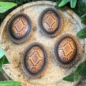 May include: Four oval-shaped, copper-colored metal embellishments with a scalloped edge and a central diamond design. The embellishments are arranged on a light-colored, textured surface with green foliage.
