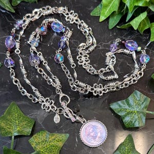 May include: A silver-toned necklace with multiple strands, featuring iridescent purple beads and a pendant with a cameo design. The necklace has intricate chain links and decorative floral elements.