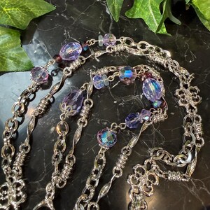 May include: A silver-toned necklace with multiple strands, adorned with iridescent and purple beads. The necklace features a mix of chain styles, including delicate links and twisted sections, with floral accents.