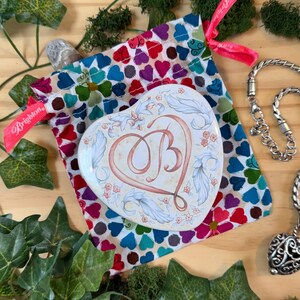 May include: A heart-shaped trinket box with the letter "B" in the center, surrounded by floral and leaf designs. The box rests on a colorful heart-patterned pouch. A silver necklace and a pink ribbon with the word "Brighton" are also visible.