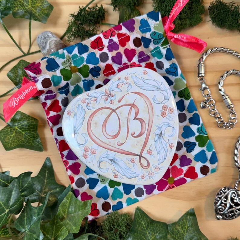 May include: A heart-shaped trinket box with the letter "B" in the center, surrounded by floral and leaf designs. The box rests on a colorful heart-patterned pouch. A silver necklace and a pink ribbon with the word "Brighton" are also visible.