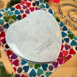 May include: A heart-shaped, light-colored jewelry box with a silver rim, resting on a colorful fabric patterned with hearts and flowers. The box has text that reads "Jewelry designed in California Packaging made in China Exclusively for BRIGHTON."