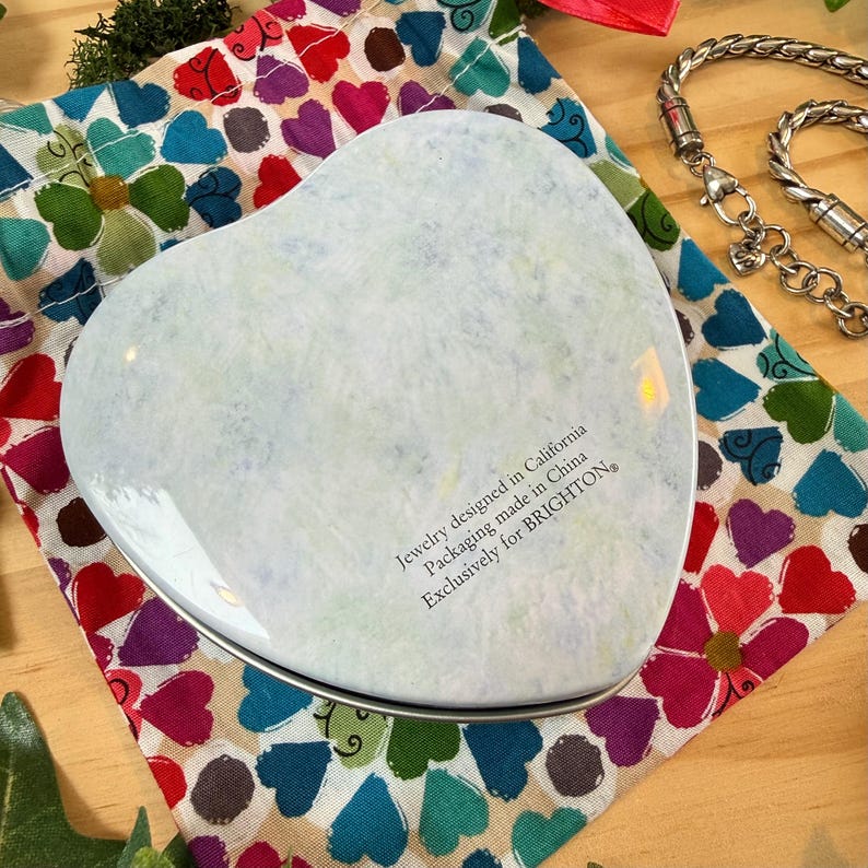 May include: A heart-shaped, light-colored jewelry box with a silver rim, resting on a colorful fabric patterned with hearts and flowers. The box has text that reads "Jewelry designed in California Packaging made in China Exclusively for BRIGHTON."