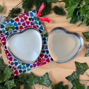 May include: Two heart-shaped metal tins, one filled with a pearlescent substance, sit beside a colorful fabric bag with a heart pattern. The bag is tied with a pink ribbon. Greenery surrounds the items on a wooden surface.