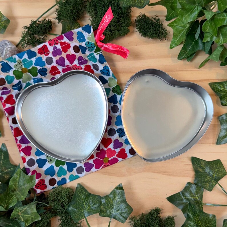 May include: Two heart-shaped metal tins, one filled with a pearlescent substance, sit beside a colorful fabric bag with a heart pattern. The bag is tied with a pink ribbon. Greenery surrounds the items on a wooden surface.