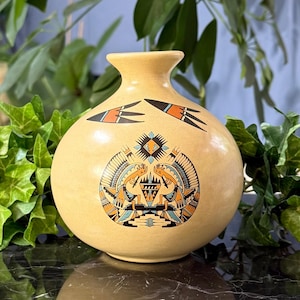 May include: A round, tan-colored ceramic vase with a narrow neck. The front features a detailed circular graphic design in black, orange, and light blue, depicting two stylized figures facing each other, with two bird-like motifs above.