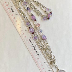 May include: Silver-toned necklace with multiple strands of chain and beaded accents in shades of purple and pink. The necklace includes a pendant with a cameo design. A ruler is placed next to the necklace.