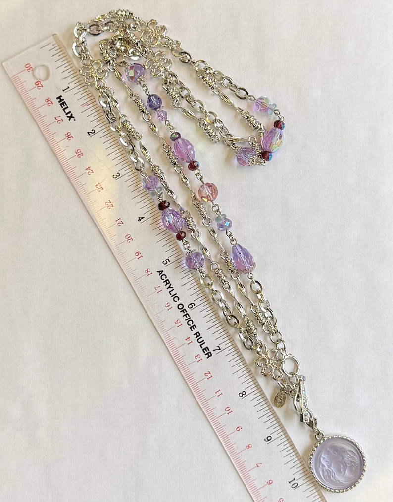May include: Silver-toned necklace with multiple strands of chain and beaded accents in shades of purple and pink. The necklace includes a pendant with a cameo design. A ruler is placed next to the necklace.
