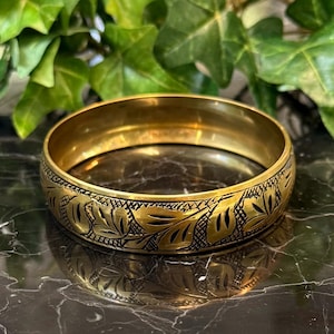 May include: A gold-toned metal bangle bracelet featuring a continuous engraved pattern of stylized leaves and cross-hatched details. The surface of the bracelet has a polished finish with dark, recessed lines highlighting the intricate floral design.