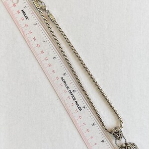 May include: Silver-toned necklace with a heart-shaped pendant. The pendant features an openwork design and is suspended from a braided chain. An acrylic ruler is placed next to the necklace, showing measurements in inches.