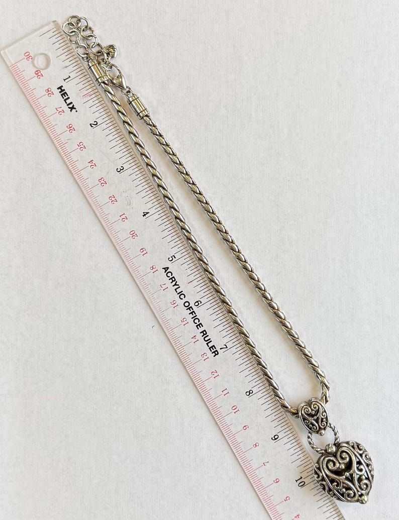 May include: Silver-toned necklace with a heart-shaped pendant. The pendant features an openwork design and is suspended from a braided chain. An acrylic ruler is placed next to the necklace, showing measurements in inches.