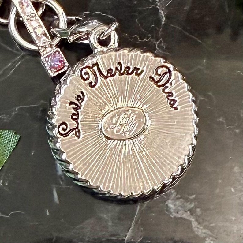 May include: A silver-toned pendant with the words "Love Never Dies" in a cursive font. The pendant has a textured, sunburst-like design in the center and a decorative border. A small, rectangular charm with pink stones is attached to the pendant.