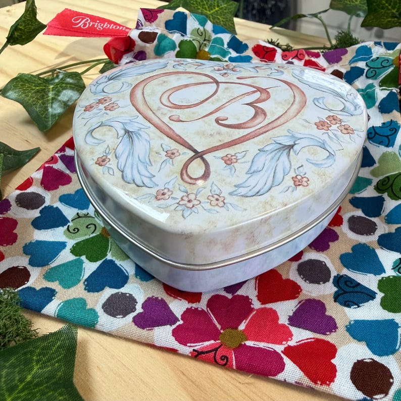 May include: A heart-shaped metal box with a floral design and the letter "B" inside a heart. The box is on a colorful fabric with heart and flower patterns. A red tag with the word "Brighton" is visible.
