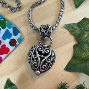 May include: A silver-toned heart pendant necklace with intricate scrollwork and a twisted rope chain. The heart-shaped pendant has a detailed openwork design. The necklace is displayed on a light wood surface with colorful fabric and green leaves.