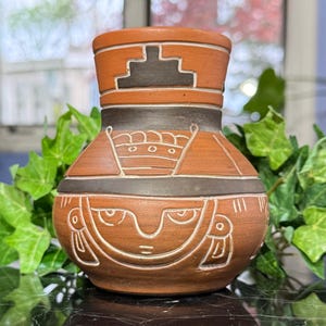 May include: A terracotta-colored ceramic vase with a dark brown band and white etched designs. The vase features geometric and stylized face motifs, with a rounded body and a flared top. The vase is approximately 6 inches tall.