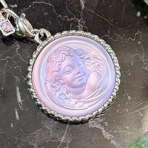 May include: A round, silver-toned pendant with a detailed relief of a woman's face. The pendant has a light purple hue and a twisted rope border. A small, square charm with a pink stone is attached to the clasp.