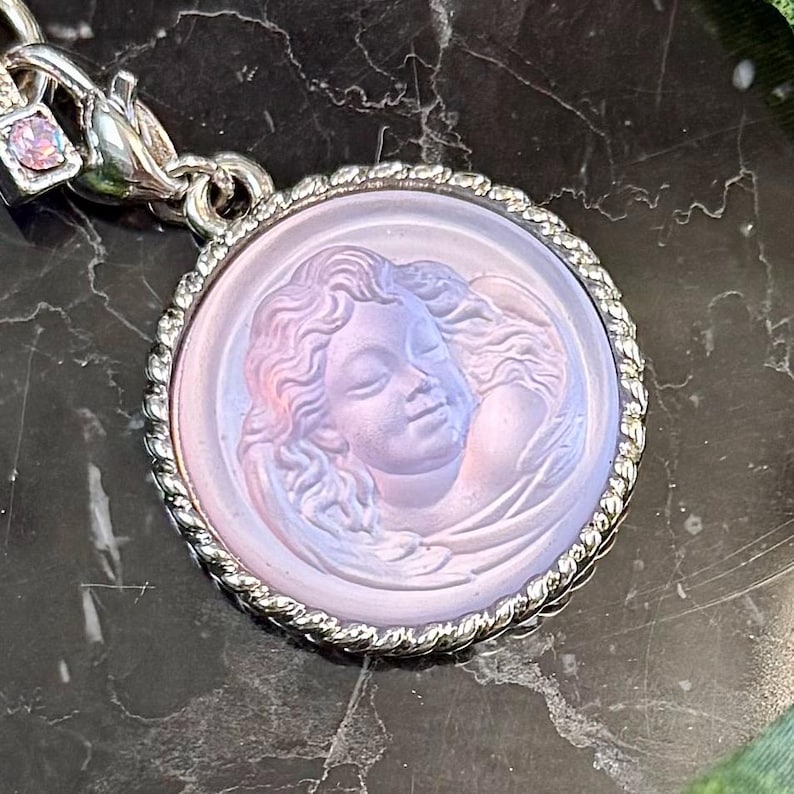 May include: A round, silver-toned pendant with a detailed relief of a woman's face. The pendant has a light purple hue and a twisted rope border. A small, square charm with a pink stone is attached to the clasp.