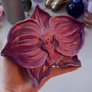 May include: A hand-painted, purple and pink orchid-shaped decorative piece. The flower has detailed petals with a glossy finish. The artwork is a unique, handcrafted item, perfect for home decor or as a gift.