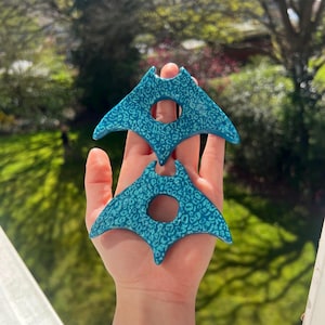 May include: Two turquoise manta ray-shaped ornaments with a leopard print pattern. Each ornament has a circular hole in the centre. The ornaments are held up in front of a window with a blurred green background.