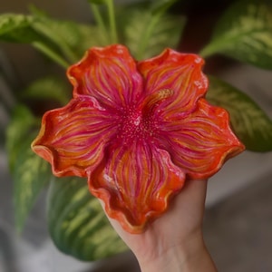 以下が含まれることがあります： 鮮やかな手描きのセラミック製の花型皿。オレンジ、ピンク、黄色の色合いで詳細なデザインが施されています。花びらは質感があり、中央は濃いピンク色です。皿は手で持たれています。