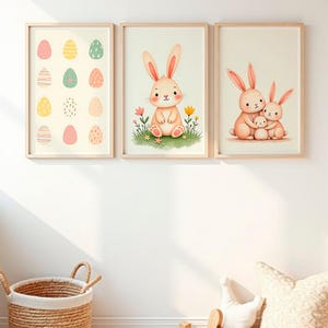 Pode incluir: Três impressões de arte emolduradas com temas de Páscoa. A impressão da esquerda mostra ovos de Páscoa coloridos. A impressão central mostra um coelhinho em um campo de flores. A impressão da direita retrata uma família de coelhos. As molduras são de madeira clara.
