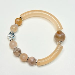 Puede incluir: Una pulsera que presenta dos tubos curvos y translúcidos de color melocotón, varias cuentas redondas de piedra en tonos beige y marrón moteado, y un pequeño dije de flor de metal plateado, unidos por espaciadores plateados.