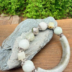 K&ouml;nnte beinhalten: Ein Jade-Armband mit einem gebogenen Jade-Teil und einer Reihe von hellgr&uuml;nen und wei&szlig;en Perlen. Silberfarbene dekorative Elemente sind zwischen den Perlen eingef&uuml;gt. Das Armband wird auf einem grauen Stein vor gr&uuml;nem Laubwerk pr&auml;sentiert.