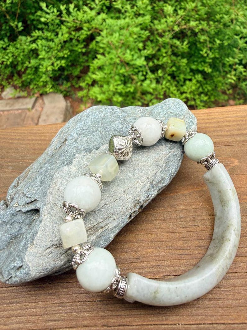 K&ouml;nnte beinhalten: Ein Jade-Armband mit einem gebogenen Jade-Teil und einer Reihe von hellgr&uuml;nen und wei&szlig;en Perlen. Silberfarbene dekorative Elemente sind zwischen den Perlen eingef&uuml;gt. Das Armband wird auf einem grauen Stein vor gr&uuml;nem Laubwerk pr&auml;sentiert.