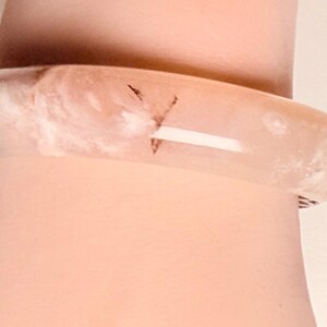 Pu&ograve; includere: Un braccialetto a cerchio traslucido rosa chiaro con aspetto marmorizzato. Il braccialetto ha una forma arrotondata e un piccolo accento rosso. Il braccialetto &egrave; indossato su un polso.