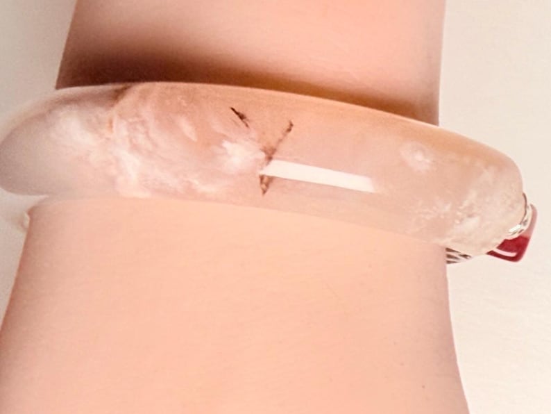 Pu&ograve; includere: Un braccialetto a cerchio traslucido rosa chiaro con aspetto marmorizzato. Il braccialetto ha una forma arrotondata e un piccolo accento rosso. Il braccialetto &egrave; indossato su un polso.