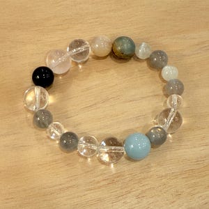 May include: A beaded bracelet featuring a variety of colorful gemstones. The bracelet includes clear, gray, black, and blue beads. The beads are strung together to form a circular shape. The bracelet is displayed on a wooden surface.