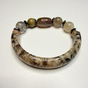 Puede incluir: Una pulsera que presenta un segmento de piedra grande, curvado y pulido con vetas en beige y marrón, unido a una serie de cuentas redondas más pequeñas en tonos bronceado, gris y marrón, con pequeñas cuentas espaciadoras metálicas oscuras.