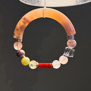 May include: A circular bracelet featuring a large, curved, translucent peach-colored segment, followed by a series of round beads in shades of pink, brown, yellow, and clear, accented with a bright red cylindrical bead and silver-toned spacers.