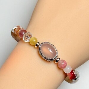 Pu&ograve; includere: Bracciale di perline con una pietra centrale ovale rosa incastonata in una cornice argentata. Il bracciale include una variet&agrave; di perline colorate nelle tonalit&agrave; del rosa, giallo e trasparente, infilate su un cinturino flessibile.