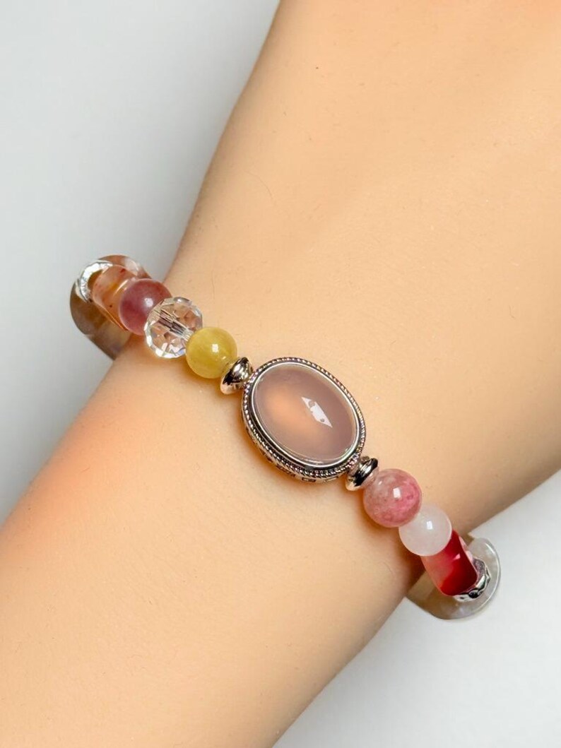 Pu&ograve; includere: Bracciale di perline con una pietra centrale ovale rosa incastonata in una cornice argentata. Il bracciale include una variet&agrave; di perline colorate nelle tonalit&agrave; del rosa, giallo e trasparente, infilate su un cinturino flessibile.